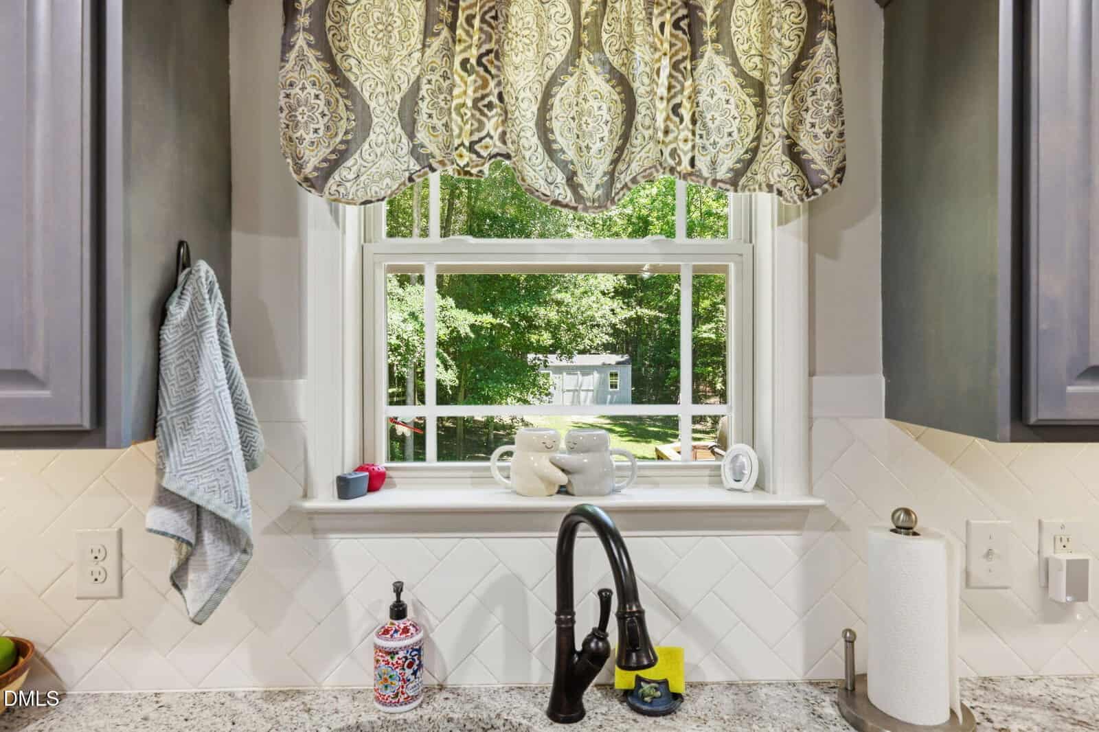 A kitchen sink with a black faucet, colorful soap dispenser, paper towels, and dish towel, beneath a window with a patterned curtain, looking out onto a green backyard with trees and a shed.