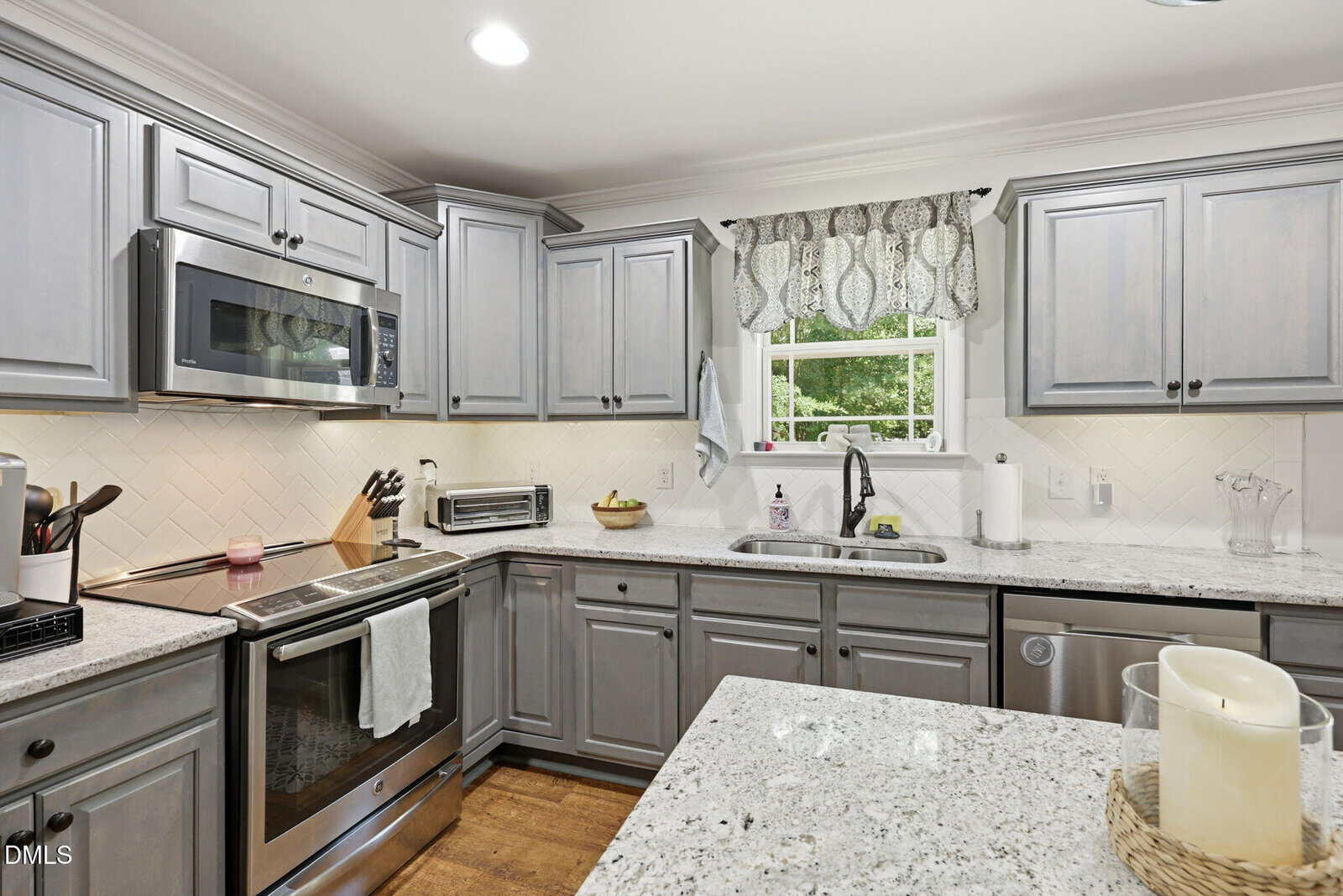 A modern kitchen with gray cabinets, stainless steel appliances, white granite countertops, a window with a patterned valance, and a kitchen island with candles.