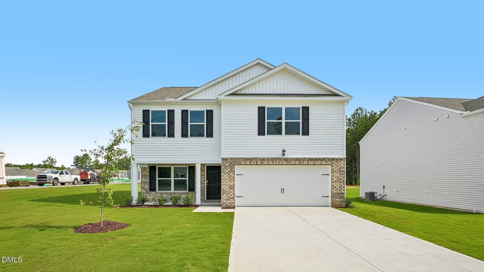 Two-story white house with black shutters, a brick accent around the garage, and a large driveway. The front yard is grassy with a small tree, and a clear blue sky is in the background.