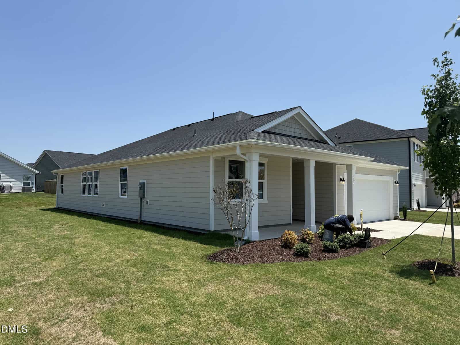 Single-story beige house with black roof, small covered front porch, attached garage, landscaped yard, and clear blue sky. A person is working near the front garden.