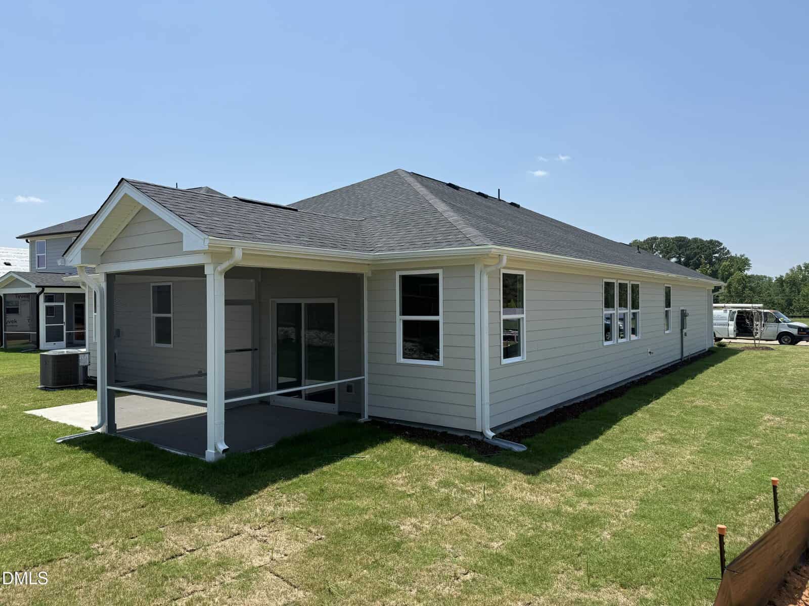 A single-story beige house with a dark roof, covered back patio, multiple windows, and a green lawn on a sunny day.