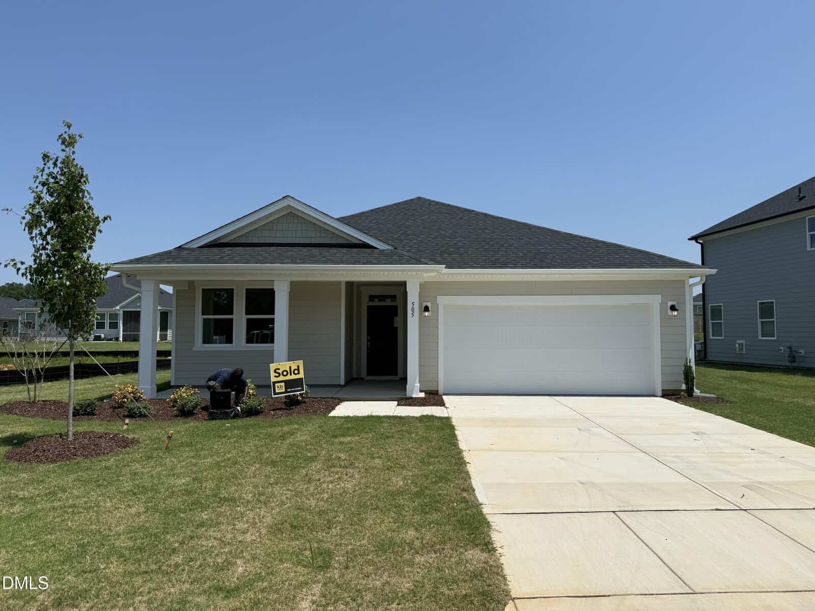 Single-story modern house with a two-car garage, front porch, and a "Sold" sign in the yard. The lawn is freshly landscaped with young trees and shrubs. Sky is clear and sunny.