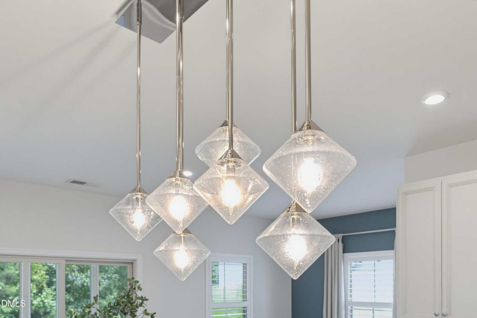 Six modern pendant lights with square, glass covers hang from the ceiling in a bright room with white walls, windows, and cabinetry visible in the background.
