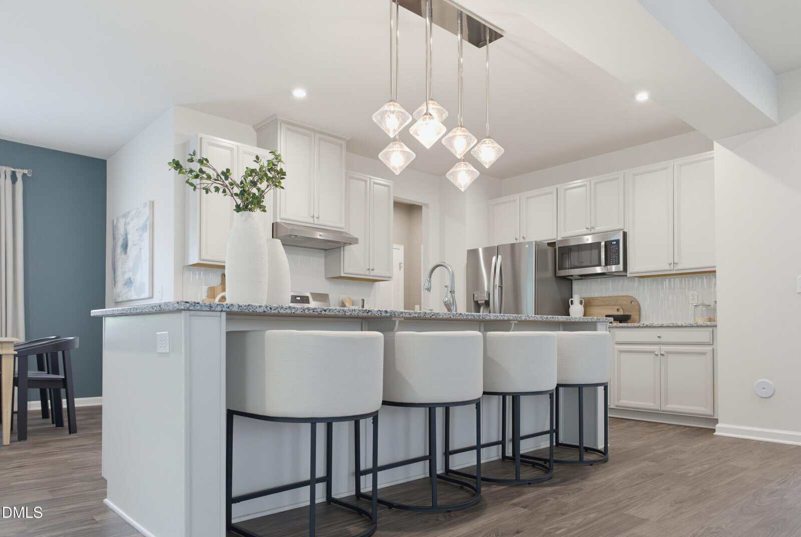 Modern kitchen with white cabinets, stainless steel appliances, a large island with granite countertop, four white cushioned barstools, pendant lights, and decorative vases with greenery. Wood flooring throughout.