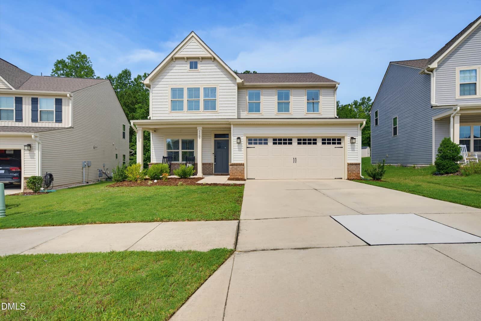 Two-story suburban house with beige siding, a covered front porch, two-car garage, and a wide driveway located at 1165 Spring Meadow Way in Wake Forest. The yard features green grass and minimal landscaping. Other similar homes are visible on each side.