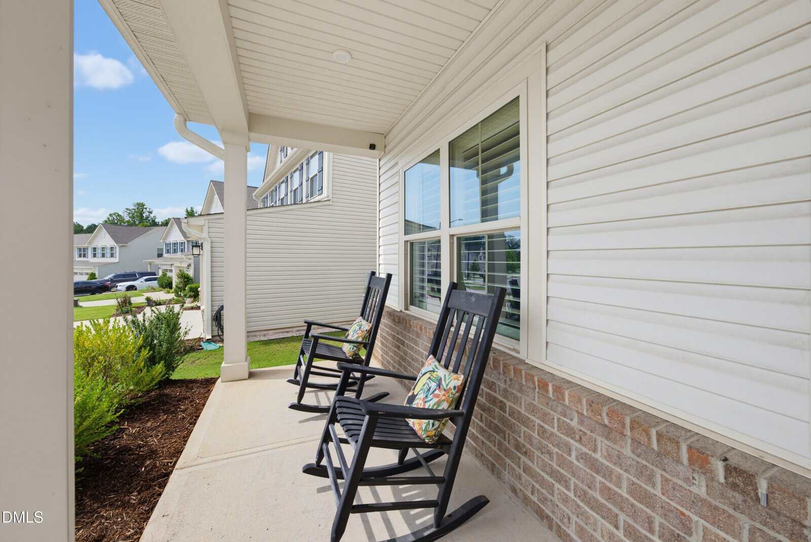 Two black rocking chairs with colorful pillows sit on a covered front porch of a house with white siding and brick trim, overlooking a neighborhood street and greenery.