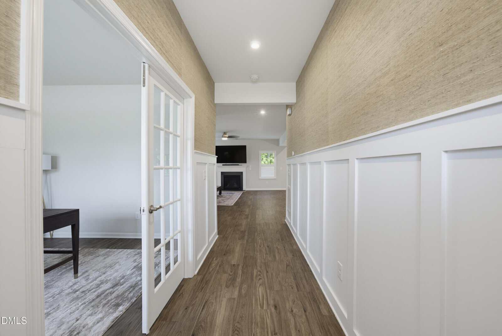 A hallway with wood floors, white wainscoting, and beige walls leads to a living room with a fireplace. Double glass doors on the left open to a bright room with a desk and lamp.