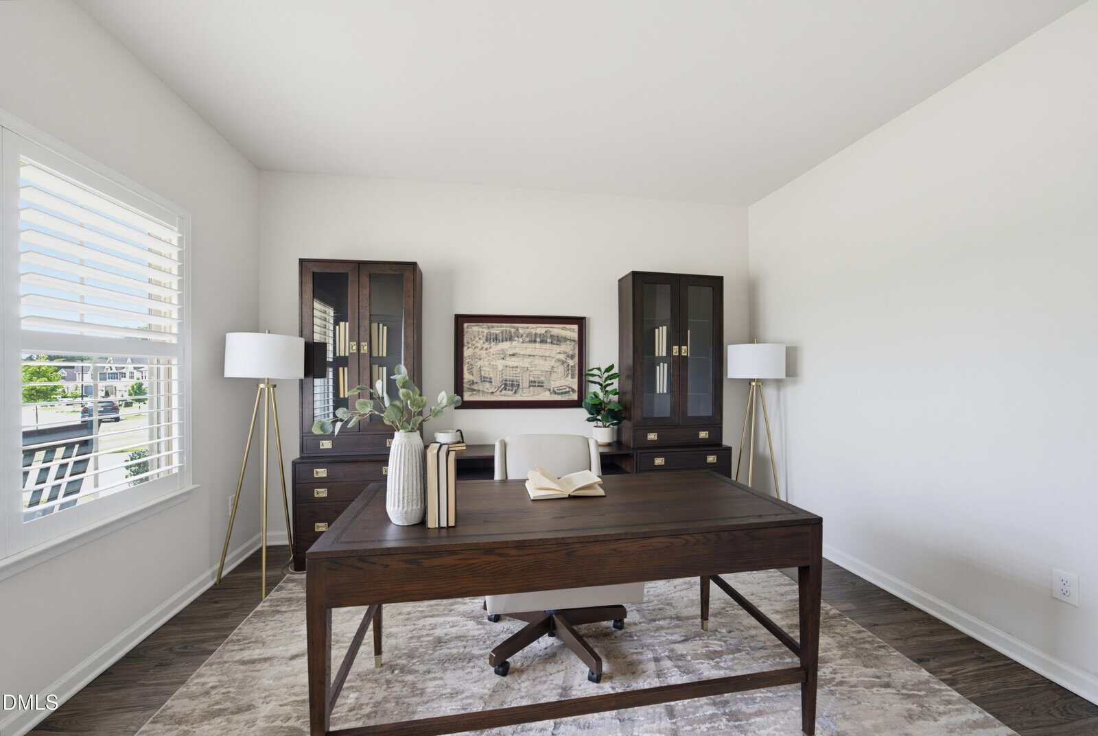 Modern home office with a dark wood desk, two glass-front cabinets, a landscape artwork, two tripod floor lamps, a potted plant, and an open book on a rug over hardwood floors. White walls and bright window light.