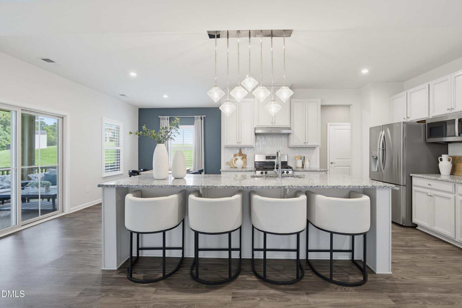Modern kitchen with a large island, five white barstools, pendant lights, stainless steel appliances, white cabinets, and a dining area with sliding glass doors to the left.