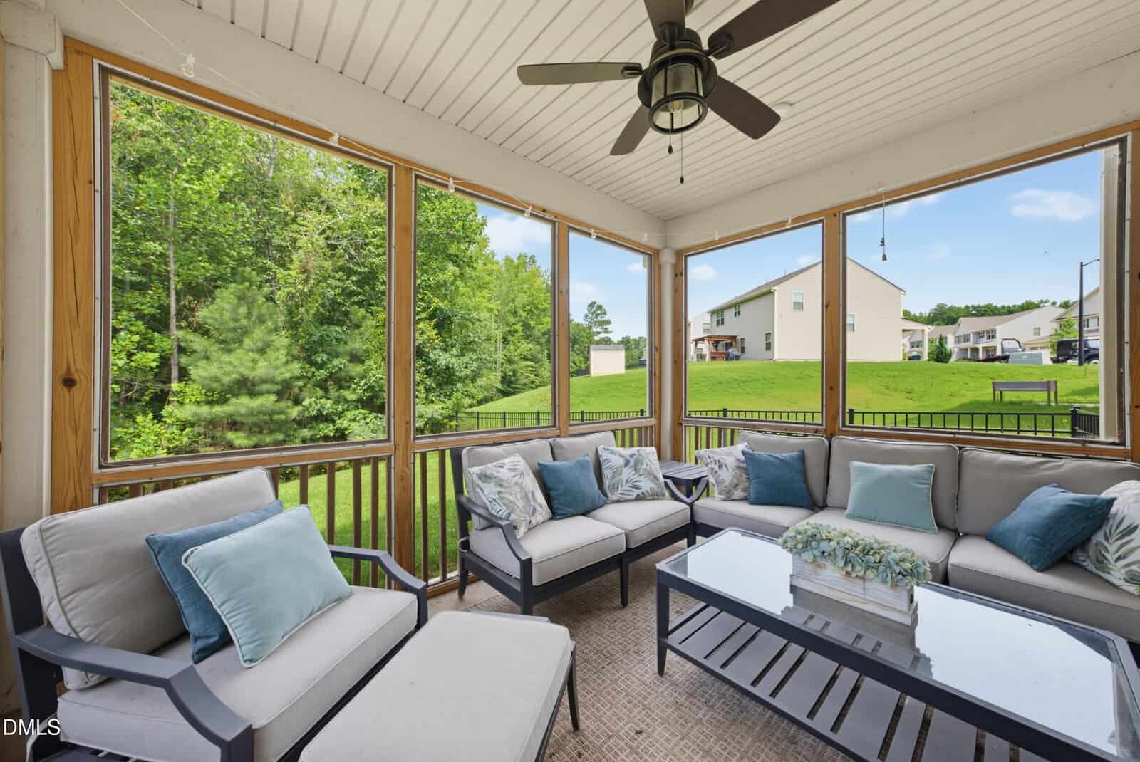 Screened-in patio with cushioned seating, a glass coffee table, decorative pillows, ceiling fan, and views of a grassy backyard with trees and neighboring houses.