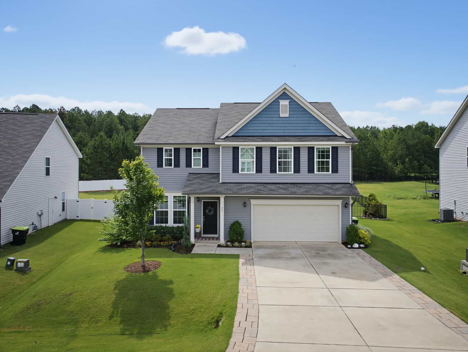Two-story blue house with white trim, a front porch, green lawn, and a two-car garage located at 75 Ashberry Lane in Franklinton. The driveway leads to the street; trees and other homes are visible in the background under a blue sky with clouds.