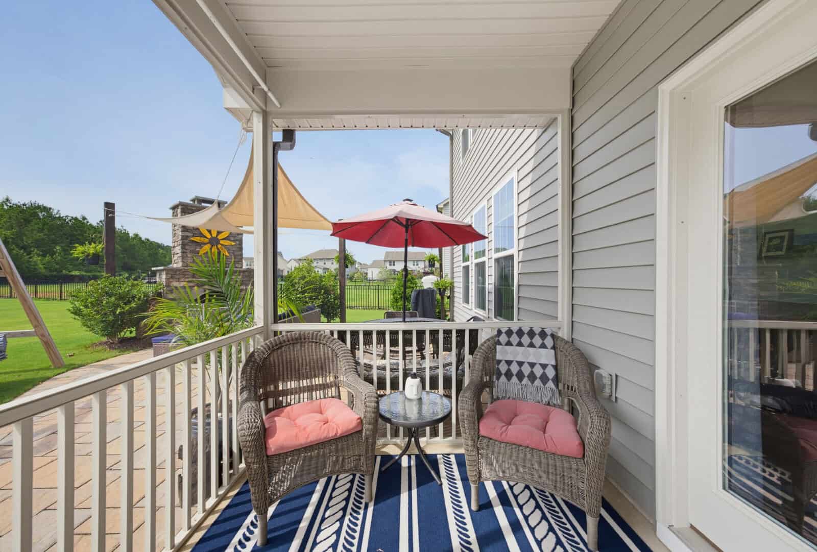 A cozy porch with two wicker chairs, a small table, decorative cushions, and a blue patterned rug; a fenced backyard, patio, and red umbrella are visible outside.
