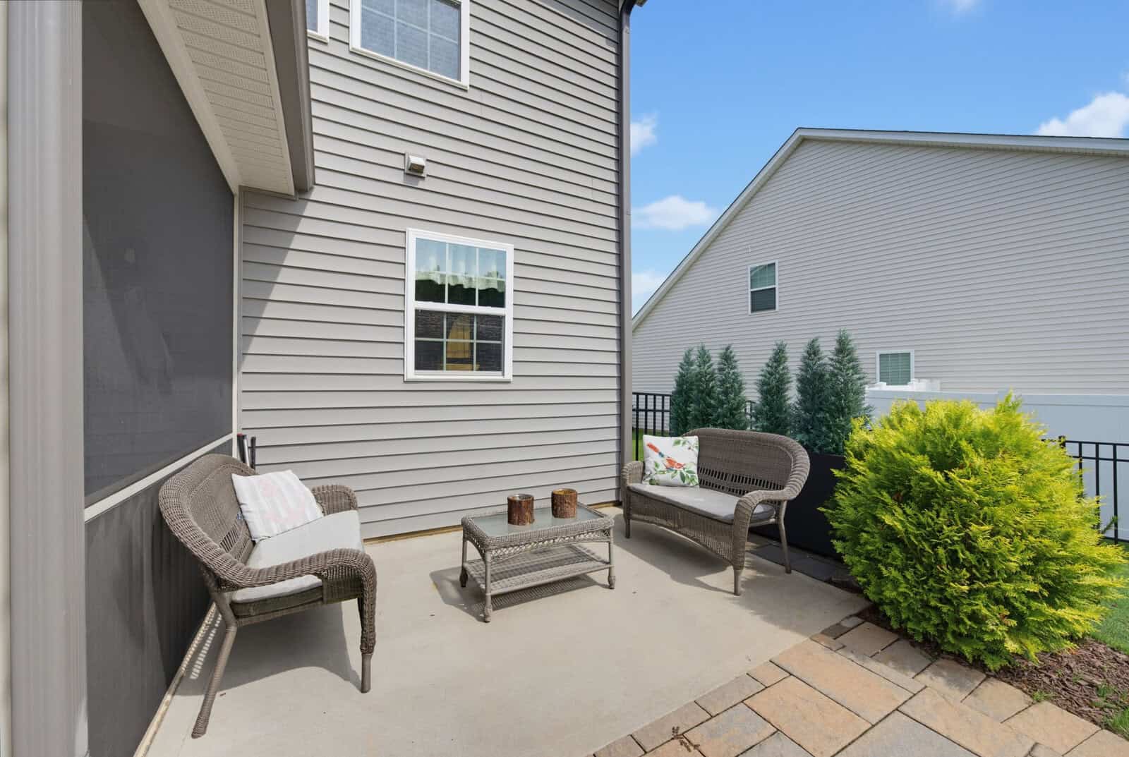 A small outdoor patio with two wicker chairs, a loveseat, a coffee table, and some cushions, next to a tan house with siding and potted plants, under a blue sky.