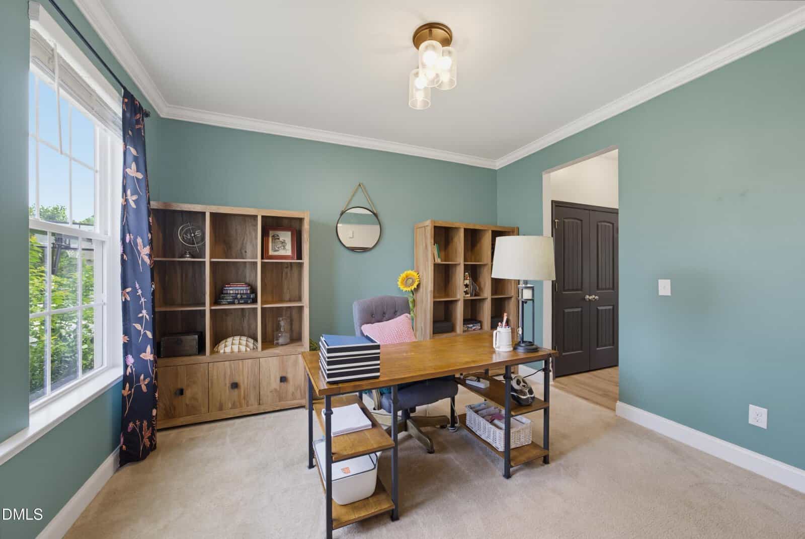 A home office with teal walls, a wooden desk, bookshelves, a gray office chair, a round mirror, and a window with dark curtains letting in natural light.