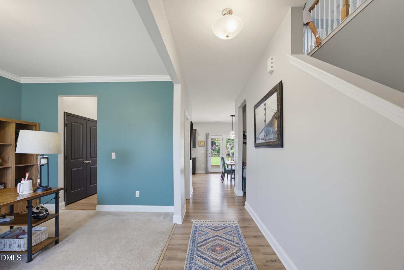 A hallway in a modern home with light walls, wood flooring, a blue accent wall, a carpeted area with a lamp and shelf, a patterned rug, and a staircase leading upstairs. Dining area visible in the background.