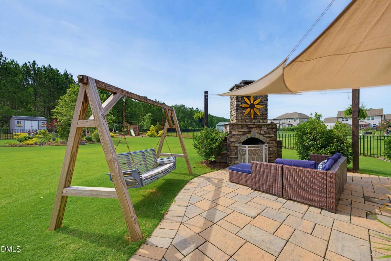 Backyard patio with wicker furniture, stone fireplace, and a wooden swing set on a green lawn. Sunshade canopy above the seating area. Houses and trees in the background on a sunny day.