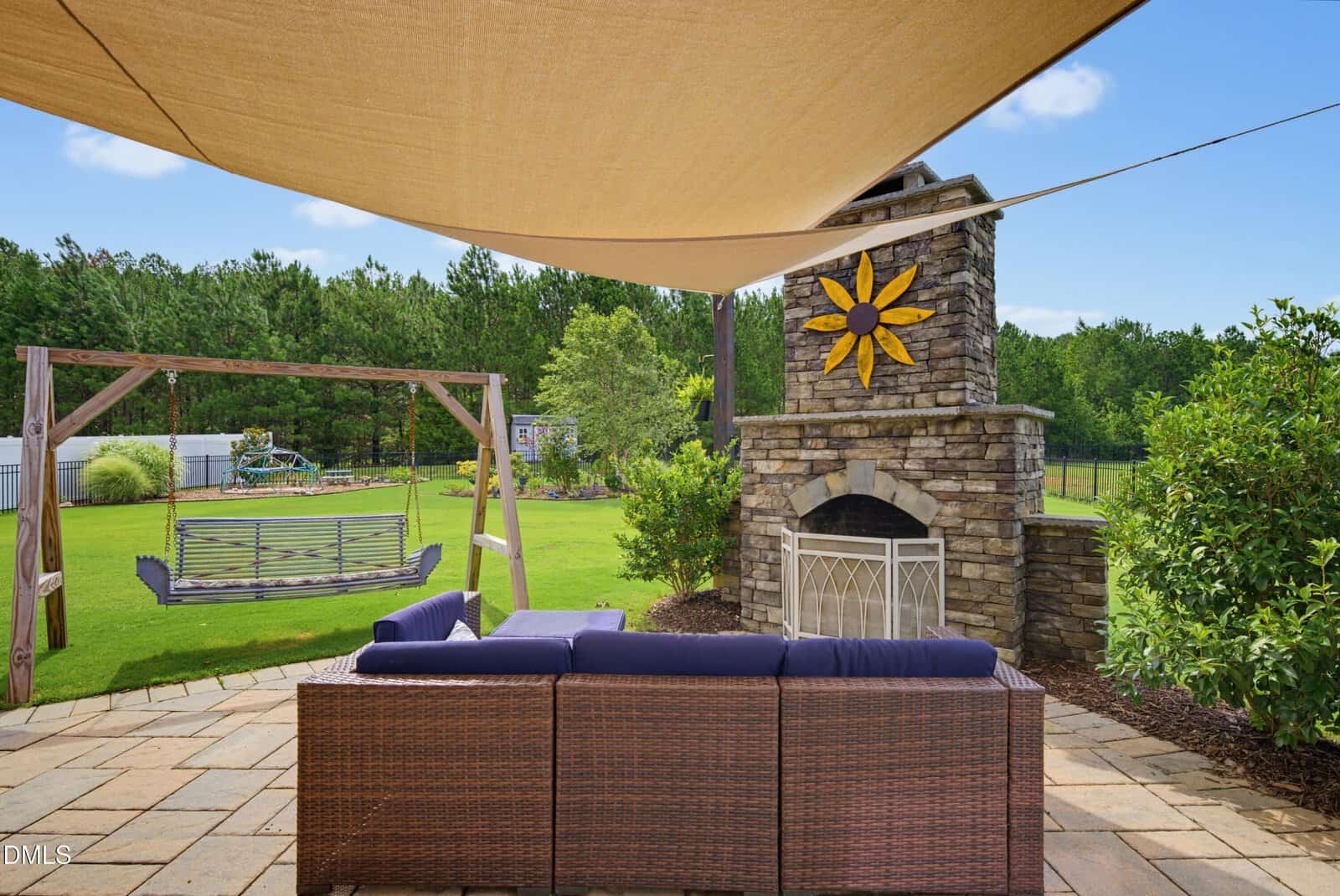 Outdoor patio with wicker sofa, stone fireplace featuring a yellow sun decoration, tan shade sail above, wooden swing, green lawn, trees, and blue sky in the background.