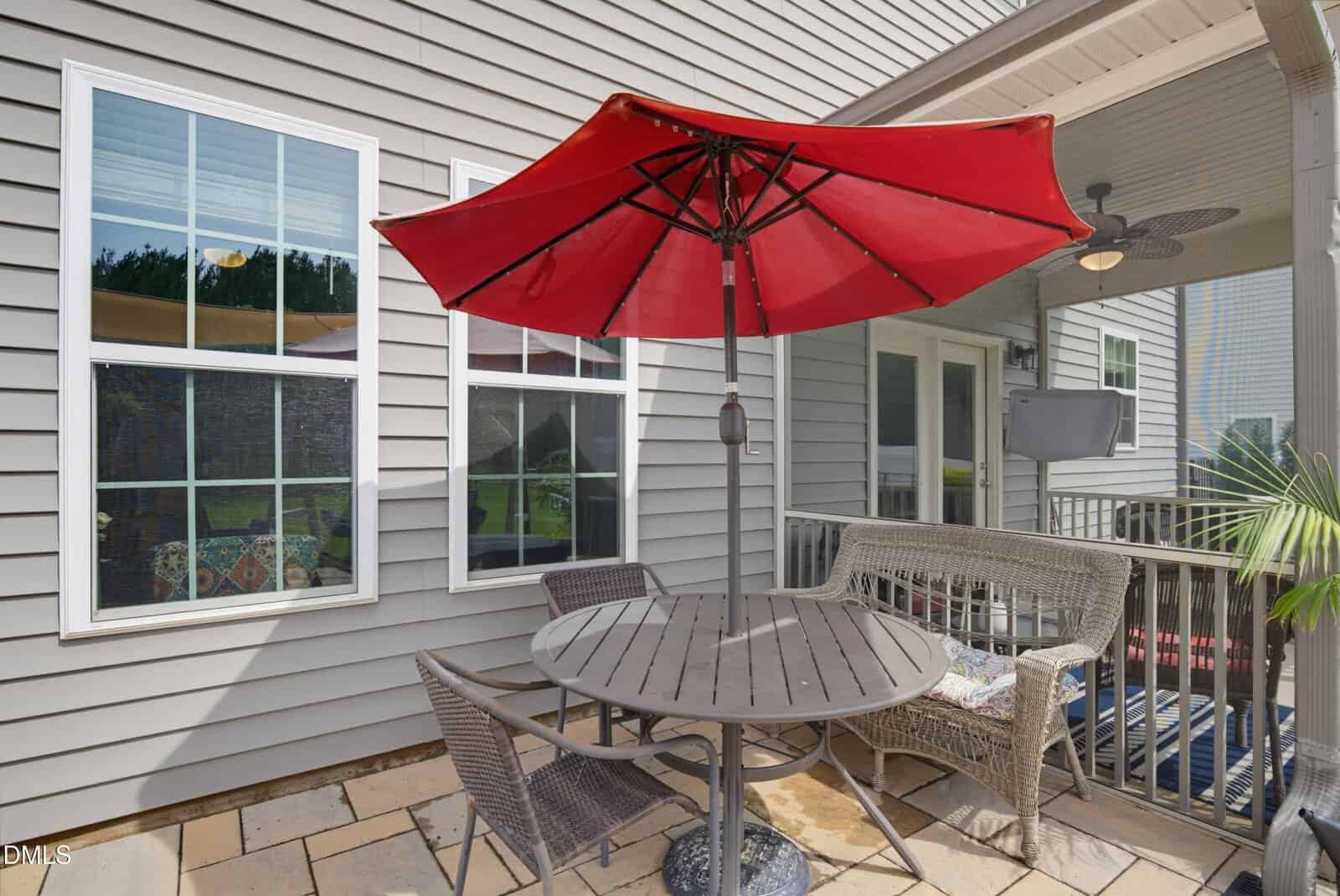 Outdoor patio with a round table, two wicker chairs, a matching love seat, and a large red umbrella. The patio is next to a gray-sided house with large windows and a door.