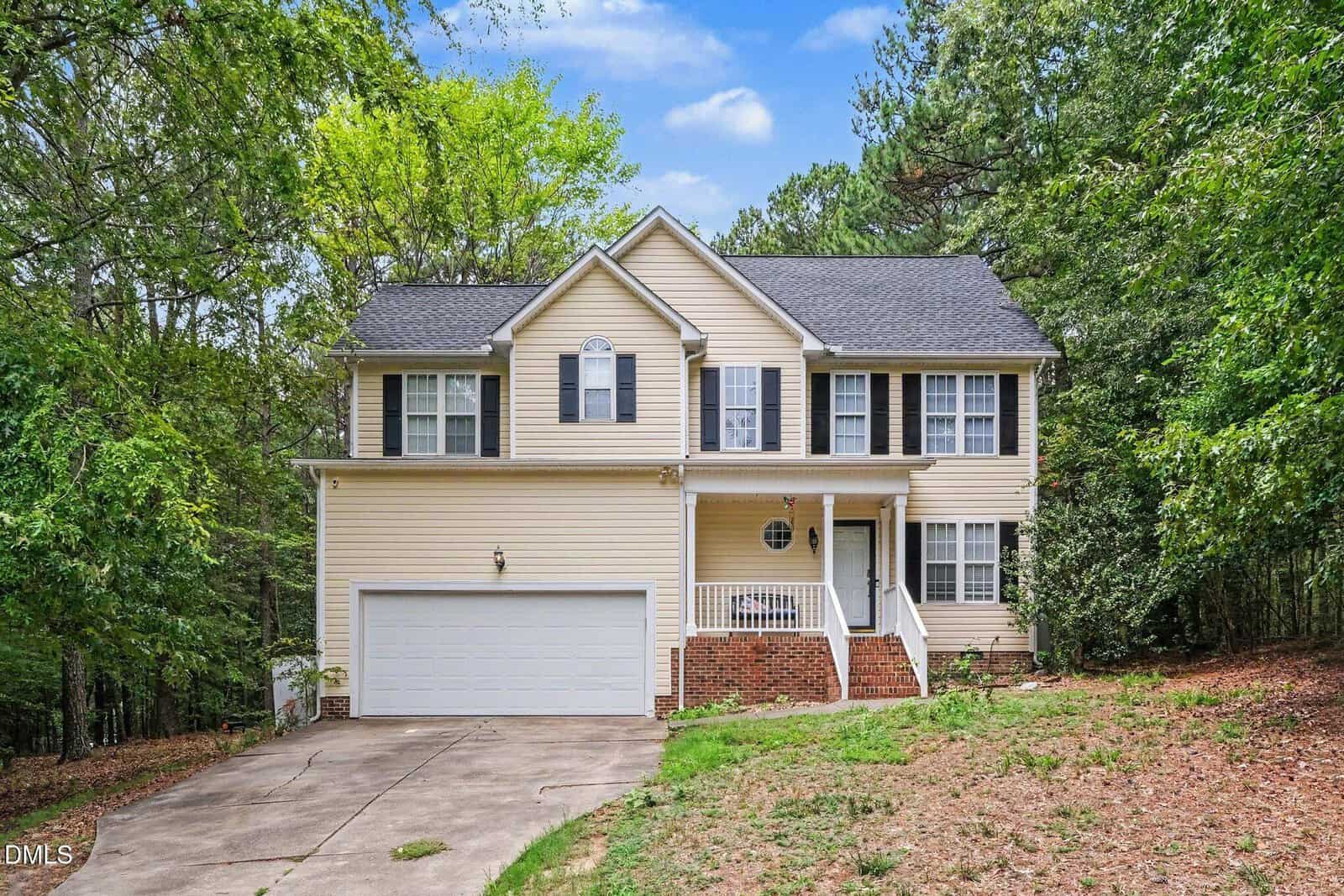 Charming two-story beige house with black shutters, white trim, a front porch, and a two-car garage located at 108 Cedar Street in Butner. Surrounded by trees, the house sits on a sloped lot with a concrete driveway and steps leading to the entrance.