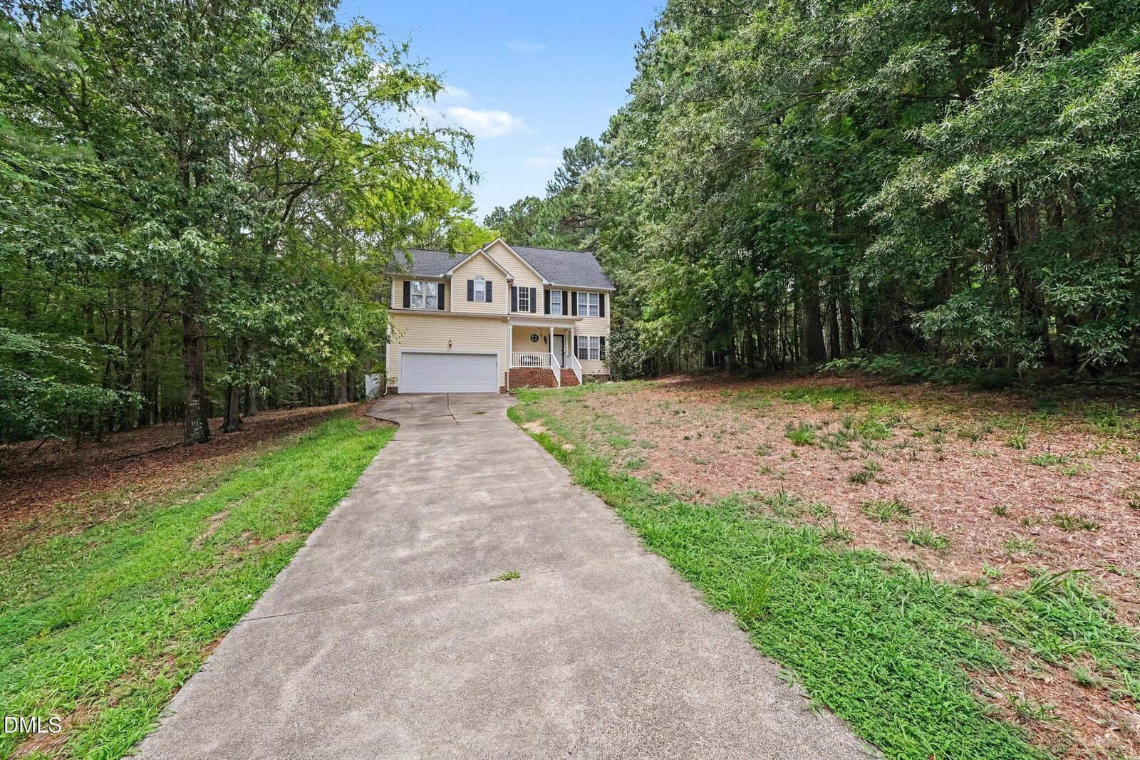A two-story beige house with white trim and a double garage sits at the end of a long concrete driveway, surrounded by trees and sparse grass on either side.