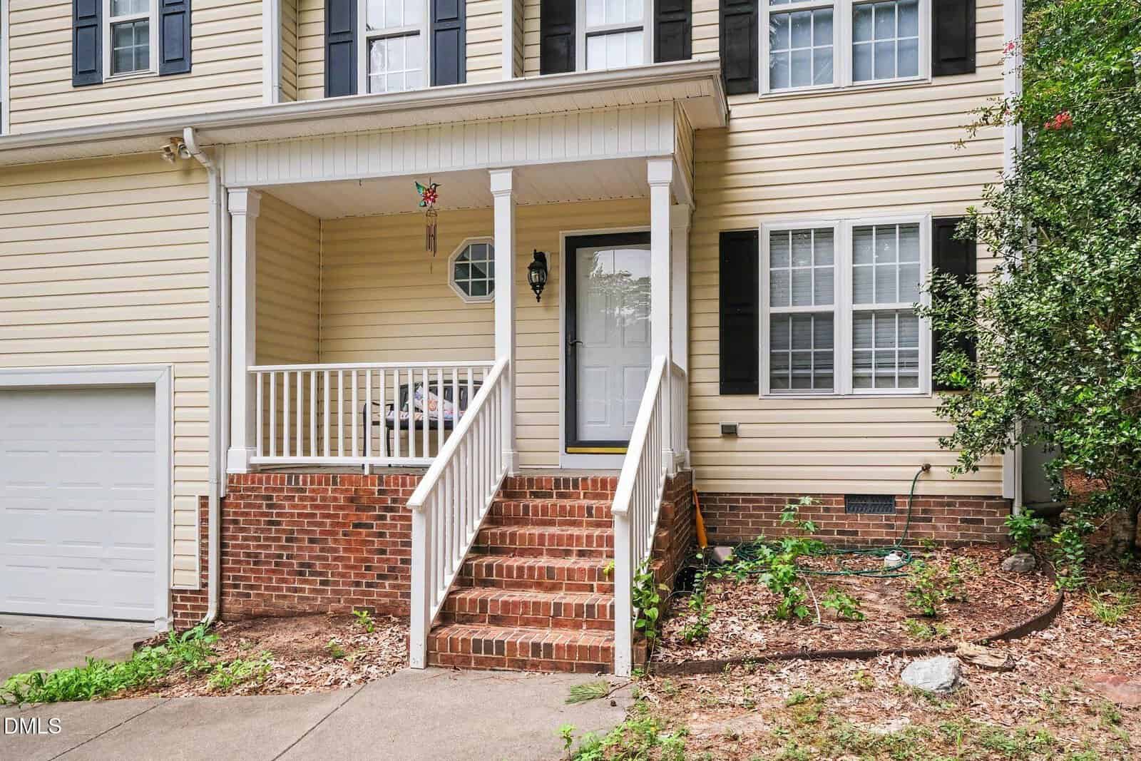 Front view of a beige house with black shutters, a white door, brick steps, a small porch with a white railing, and a garage door on the left. Some greenery grows near the steps and along the right side.