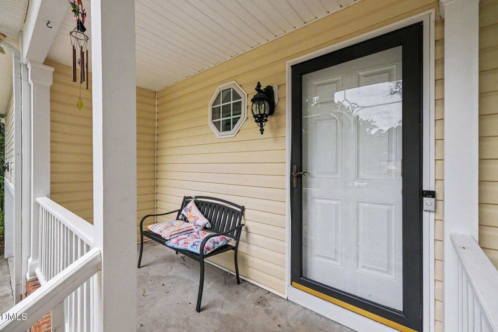 A small front porch with yellow siding, a black bench with colorful pillows, a white door with black trim, a lantern-style light, wind chimes, and white railing.