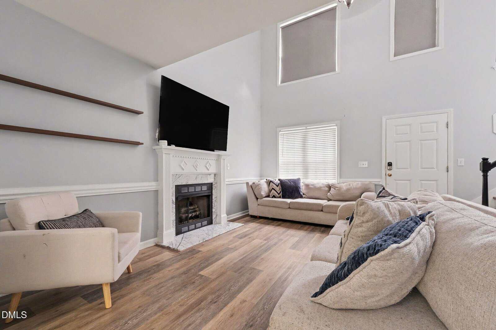 Modern living room with light gray walls, wood flooring, beige sofas, a white fireplace, large mounted TV, empty wall shelves, tall windows, and a closed white door. Natural light fills the space.