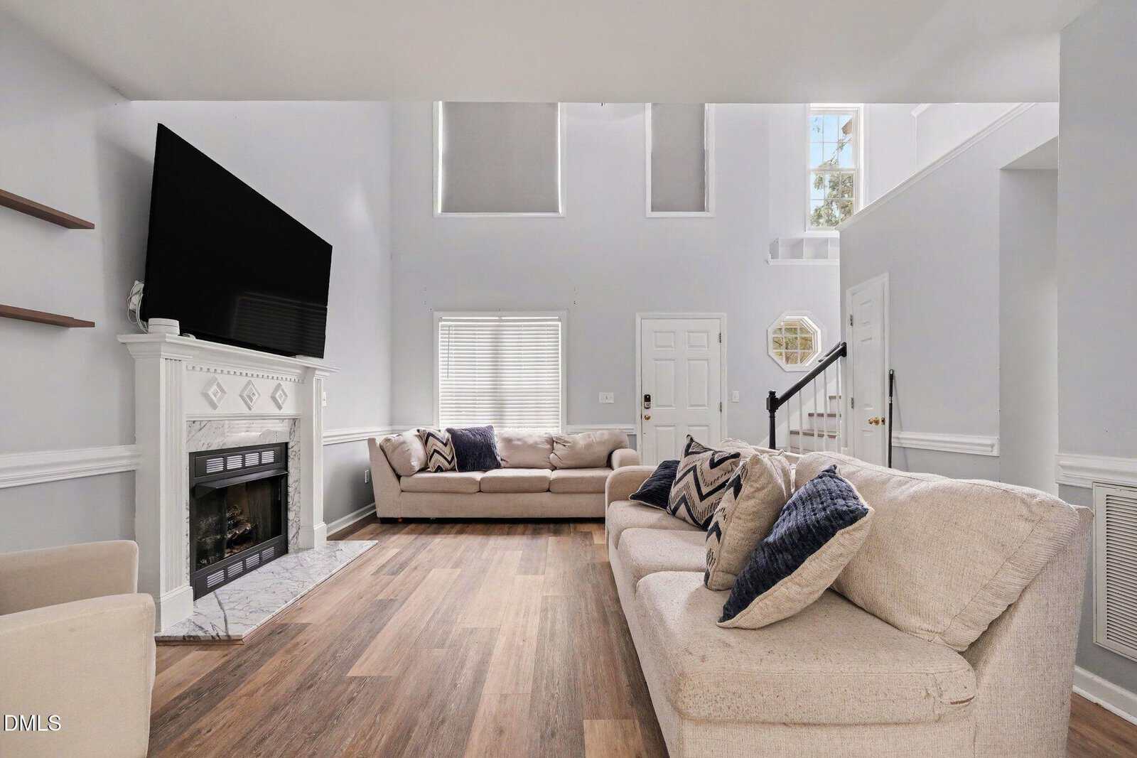 Modern living room with light gray walls, beige sofas, patterned pillows, a fireplace with a large TV above, wood flooring, and large windows letting in natural light. A staircase is visible near the front door.