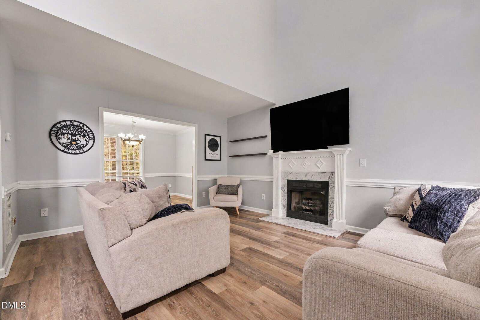 Modern living room with beige sofas, a fireplace with a large TV above, wood floors, light gray walls, and two armchairs. Adjacent dining area visible through an open doorway, lit by a chandelier.