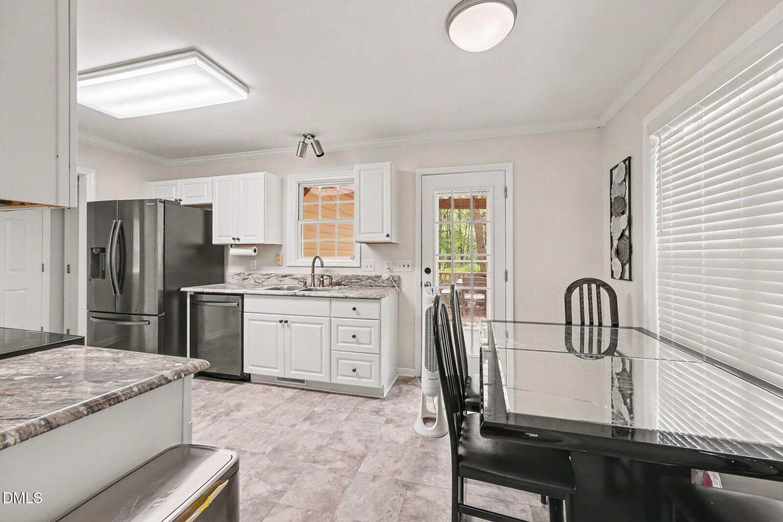 Bright kitchen with white cabinets, stainless steel appliances, a marble countertop, glass dining table with black chairs, and a door leading outside. Large window with blinds lets in natural light.