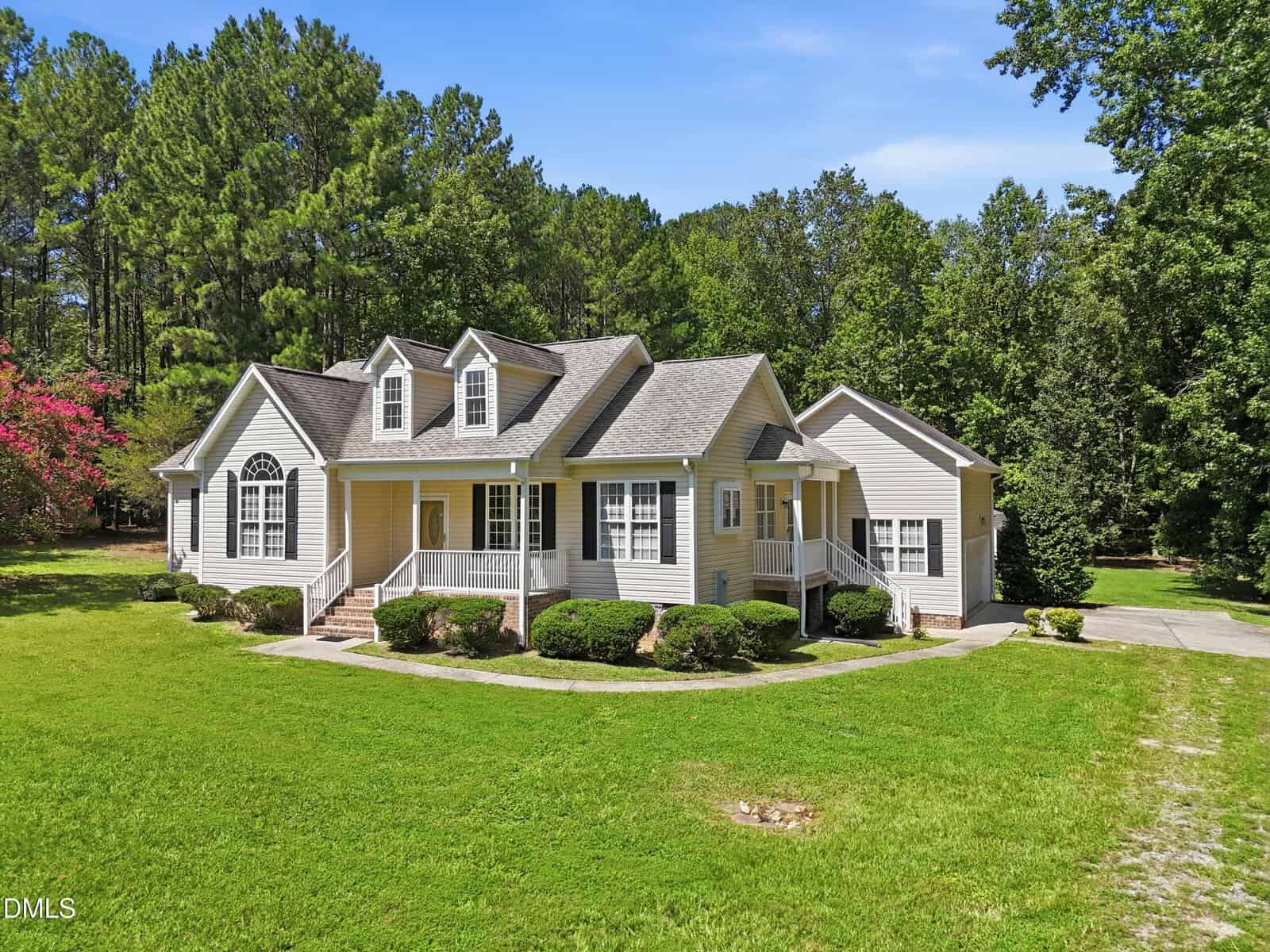 A light gray, single-story house with white trim and dormer windows has a front porch with steps, surrounded by green lawn and trees under a clear blue sky. A driveway is visible to the right of the house.