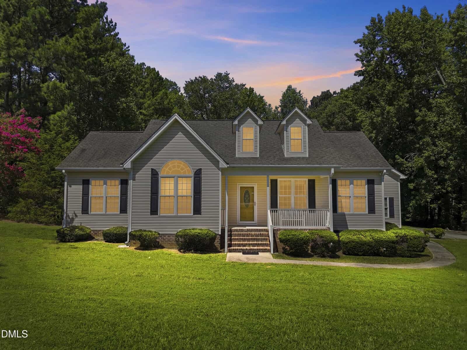 A gray single-story house with white trim, dormer windows, and a front porch sits on a well-manicured lawn with shrubs. Trees surround the property, and the sky shows a sunset with blue and orange hues.