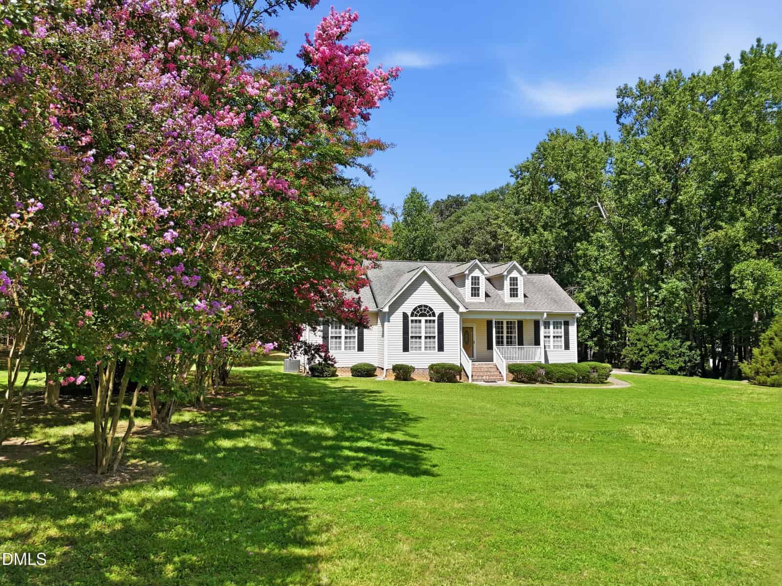 A white house with dormer windows sits on a large, green lawn bordered by trees. Pink flowering trees are on the left, and the sky is clear and blue. The setting appears peaceful and well-maintained.