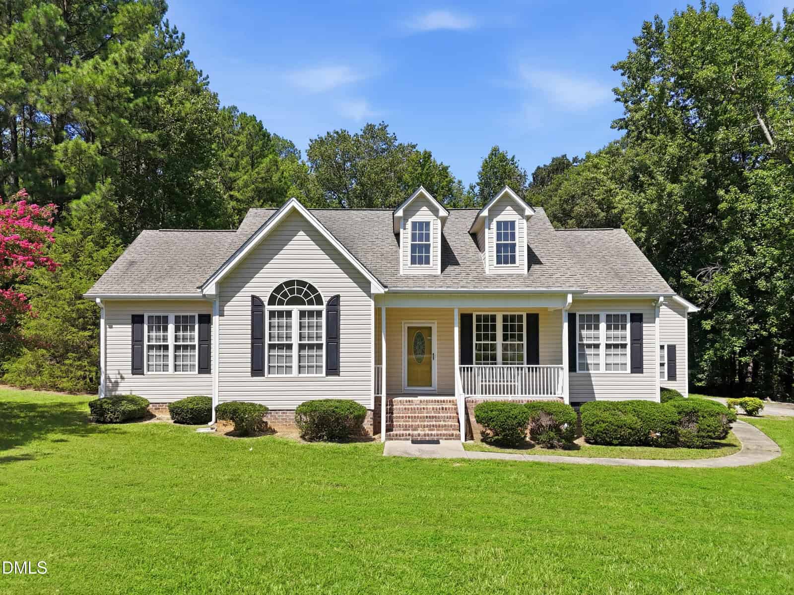 Single-story house with gray siding, white trim, and gabled roof. There are two dormer windows, a front porch, and brick steps. The yard has green grass, shrubs, and trees in the background under a clear blue sky.