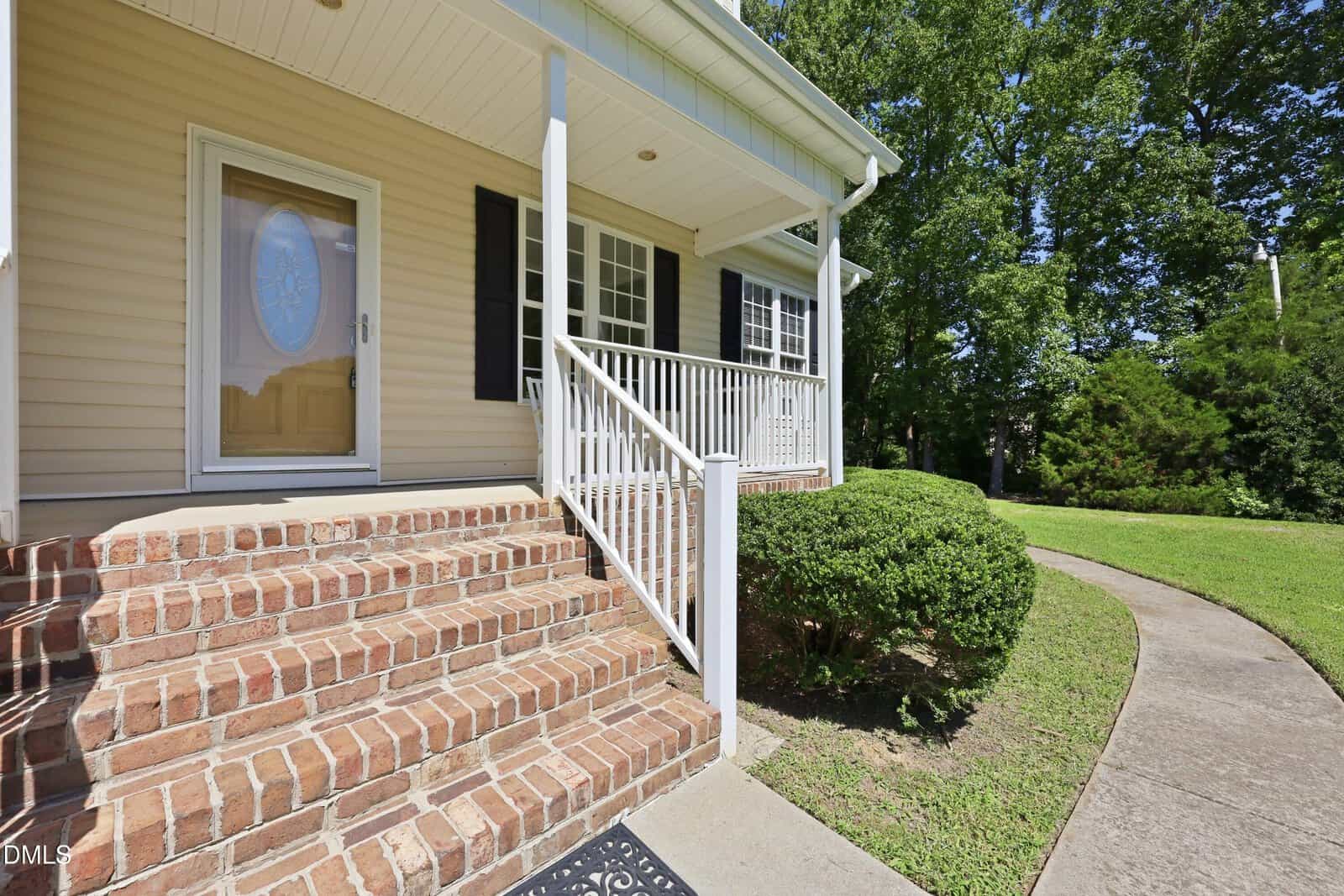 Brick steps lead to a covered front porch with white railings and beige siding. A curved sidewalk runs beside trimmed bushes and a green lawn with trees in the background.