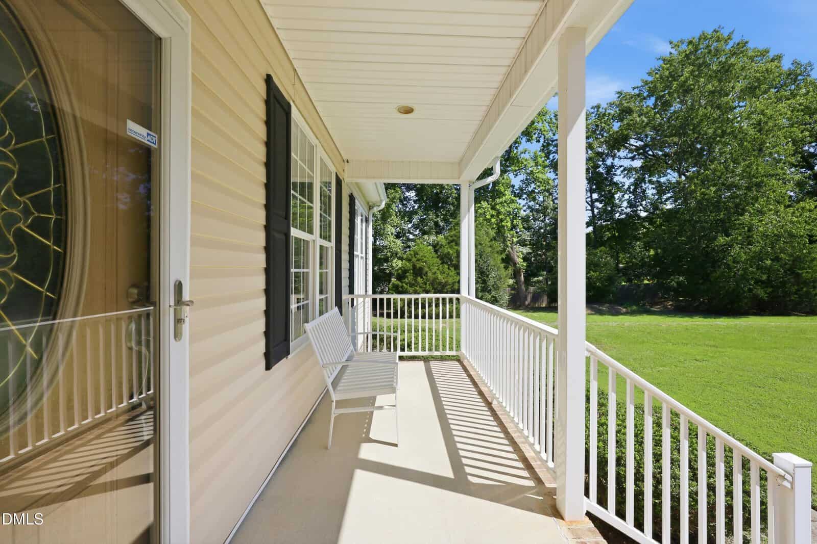 A white porch with a bench, white railing, beige siding, and black shutters, overlooking a green lawn and trees on a sunny day.