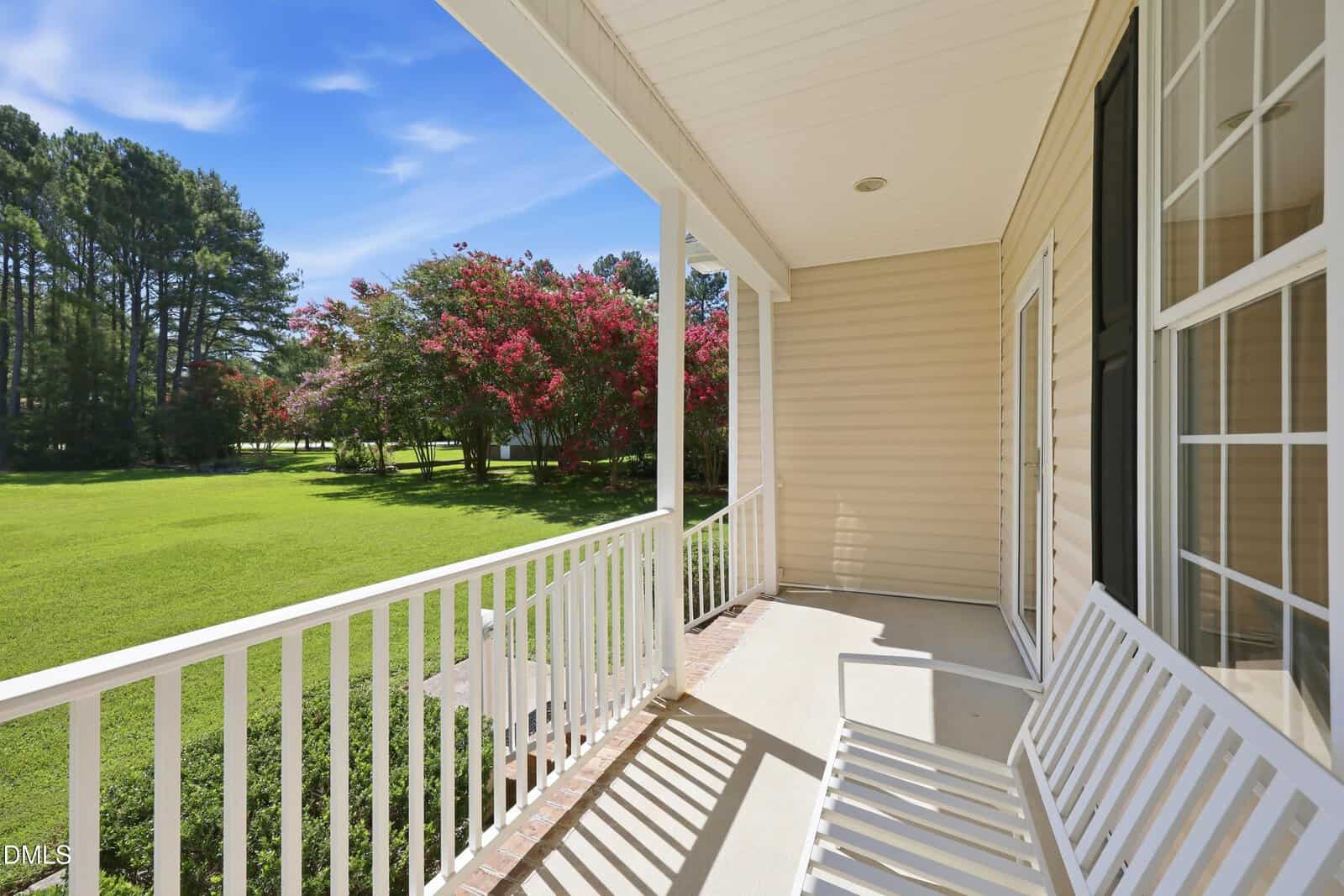 A white bench sits on a covered porch with white railings, overlooking a green lawn, trees, and flowering bushes under a clear blue sky.