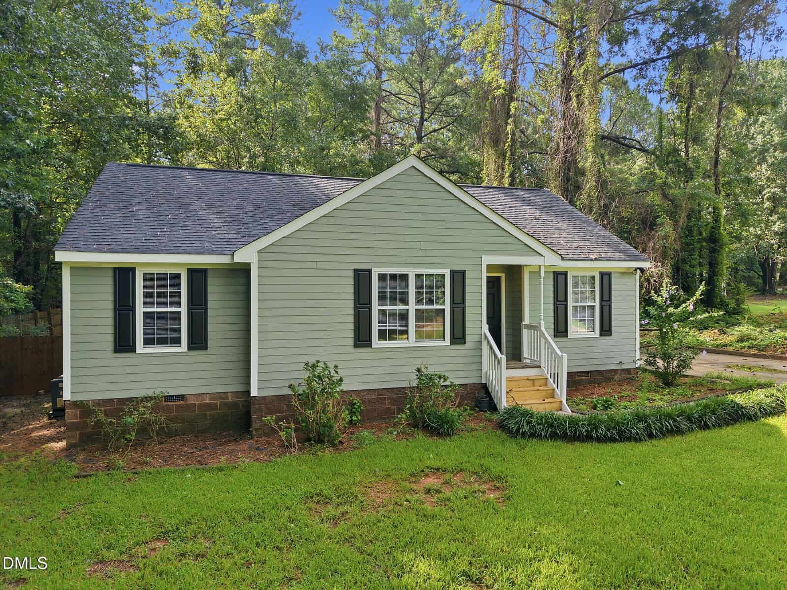 A small, single-story green house with white trim and black shutters sits on a grassy lawn, surrounded by tall trees. A front porch with white railings leads to the entrance.