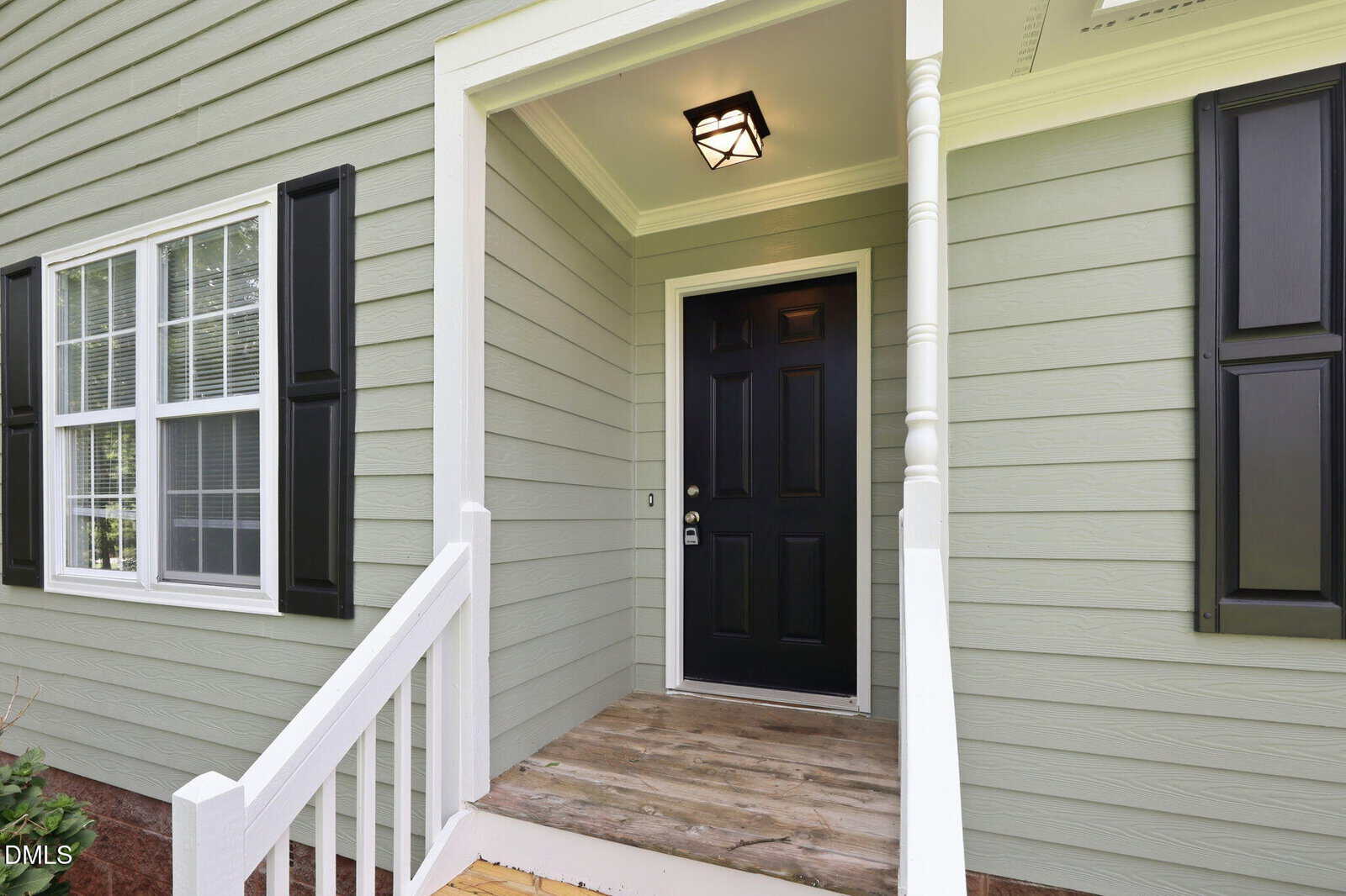 A small porch with white railings and light green siding leads to a black front door. A modern light fixture hangs above, and two windows with black shutters are on the left side of the entrance.