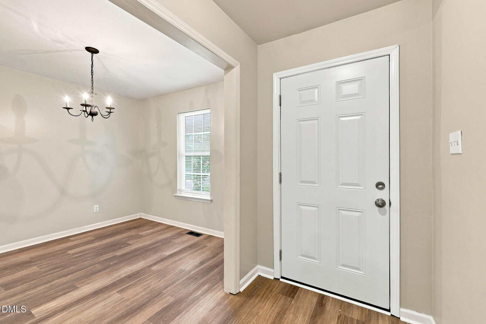 A white front door opens to a room with wood flooring and beige walls; an adjacent dining area features a window with blinds and a black chandelier casting clear shadows.
