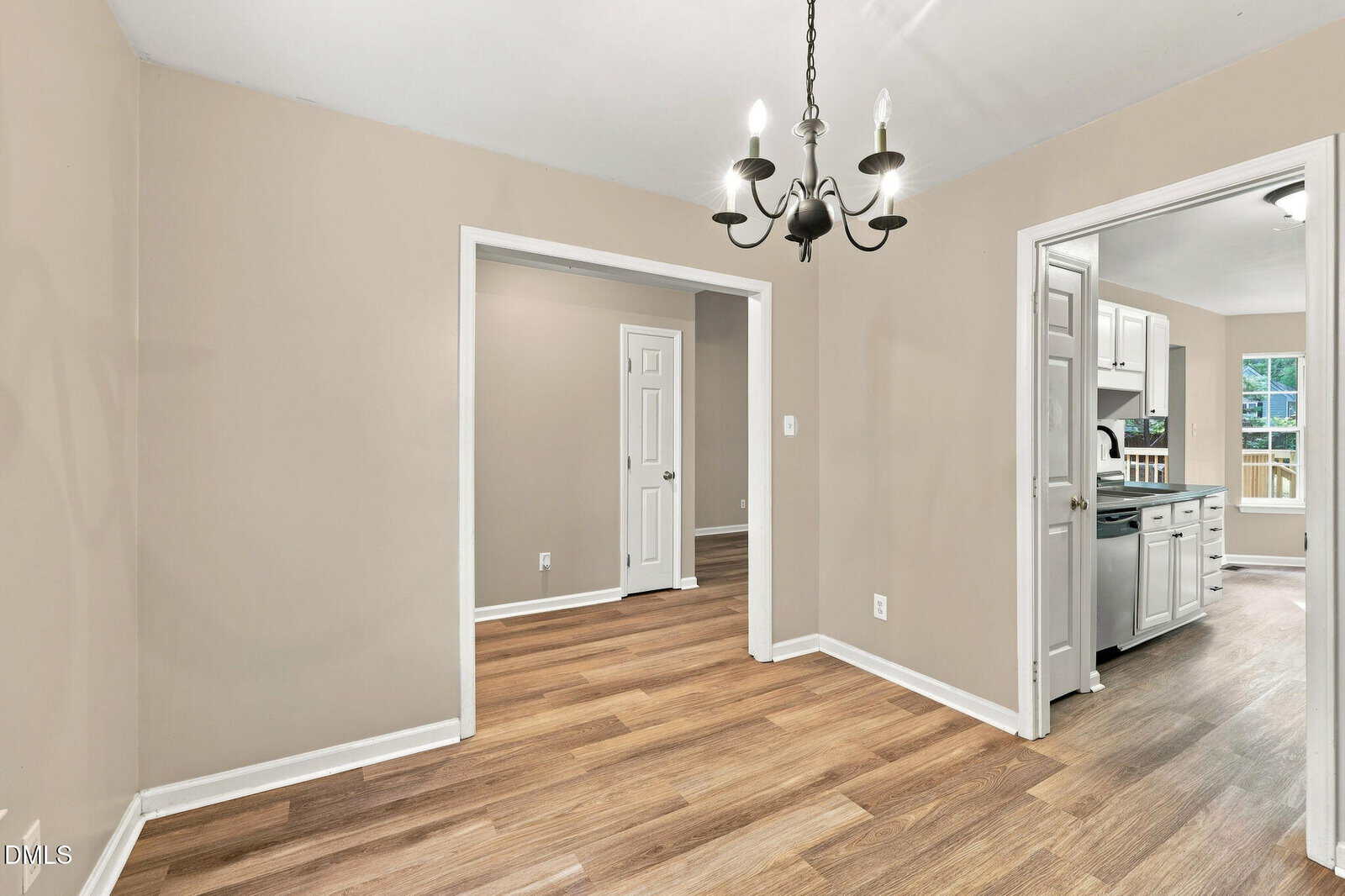 Dining room with wood flooring, beige walls, and a black chandelier. Two doorways lead to other rooms, one showing a kitchen with white cabinets and another with a closed white door.