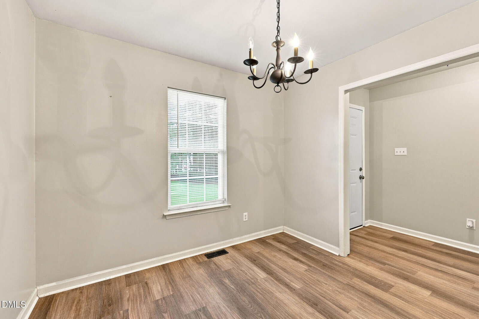 Empty room with light gray walls, wood flooring, a single window with blinds, and a black chandelier casting shadows on the wall; doorway leads to another area with similar flooring and a white door.