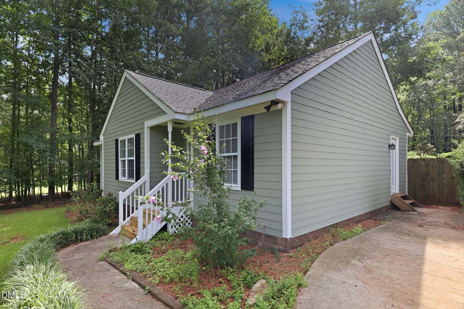 Light green single-story house with white trim and a small front porch, surrounded by trees and a lawn. Concrete path leads to the front steps; a flower bush grows near the entrance.