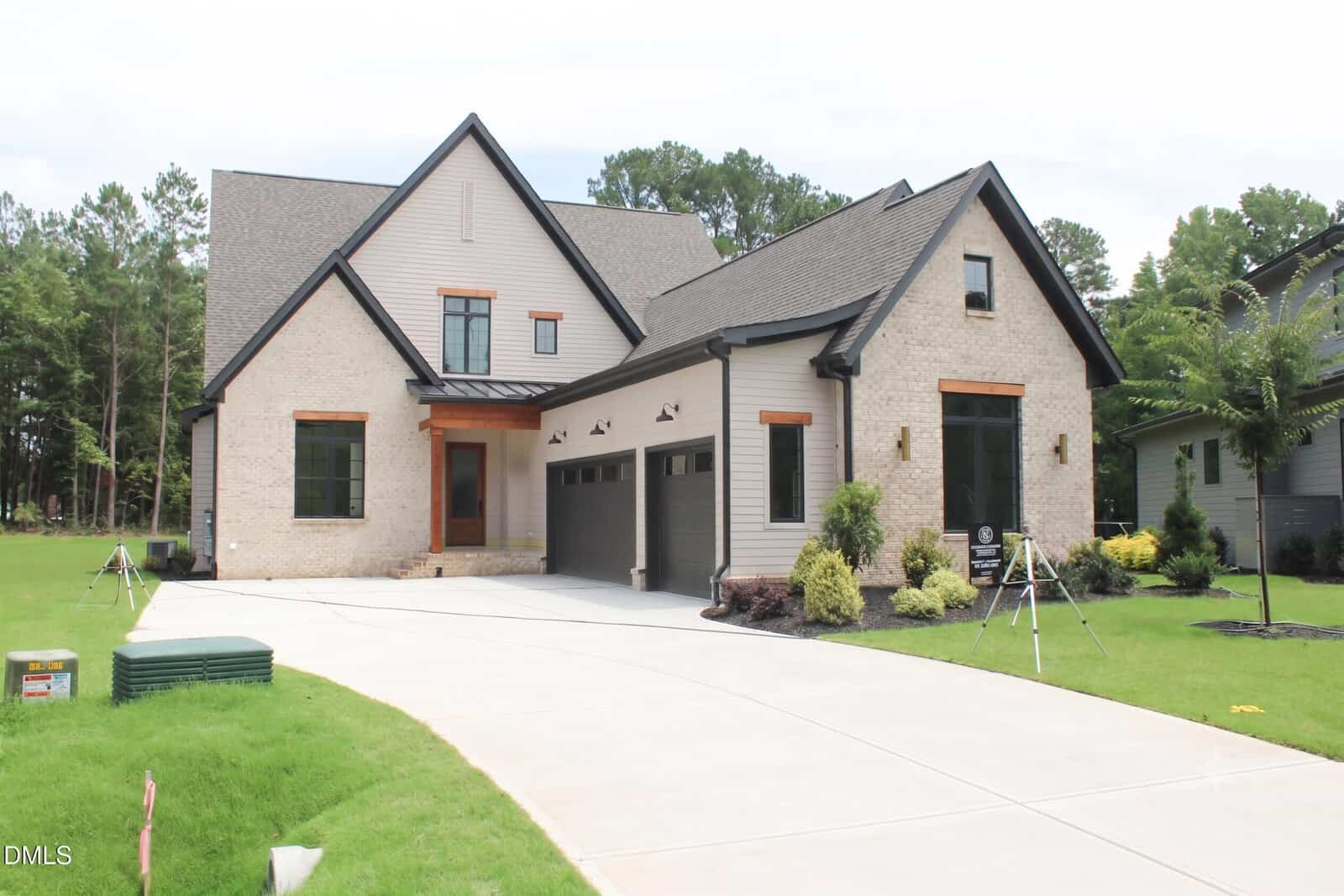 Modern two-story house with light brick and gray siding, three-car garage, and a clean, wide driveway located at 15 Geranium Drive in Youngsville. The yard is landscaped with green grass, bushes, and small trees. The home is surrounded by woods.