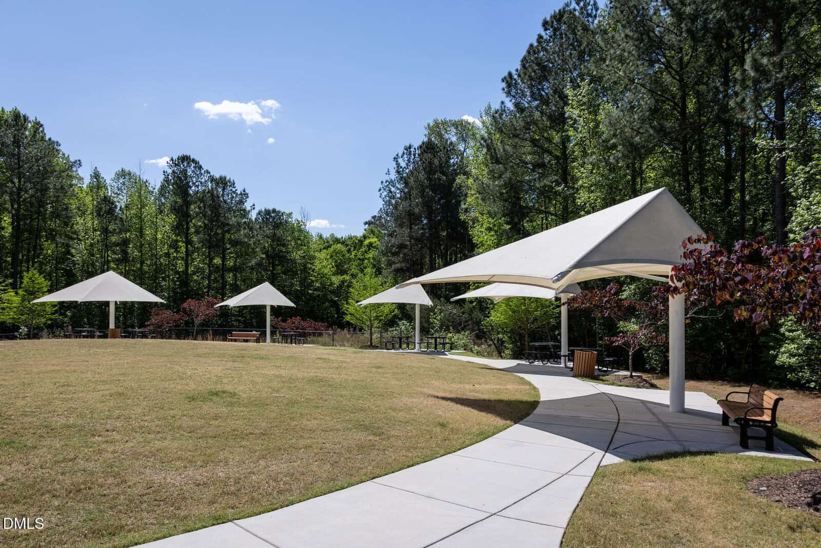A curved sidewalk leads to three white canopy-covered picnic areas on a grassy lawn, surrounded by green trees under a clear blue sky.