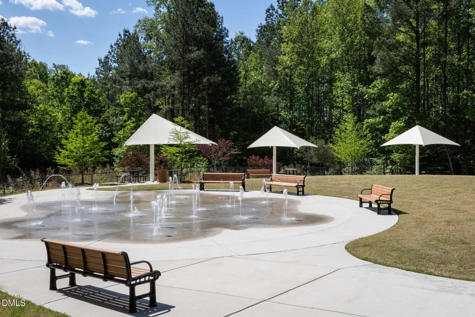 A circular splash pad with water jets, surrounded by benches and white shade structures, set in a grassy area with trees in the background under a blue sky.