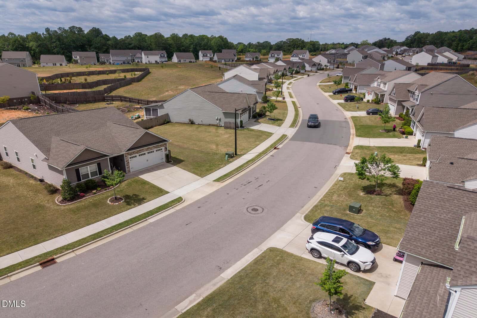 A suburban neighborhood with single-story houses, green lawns, and parked cars along a curving street under a partly cloudy sky.