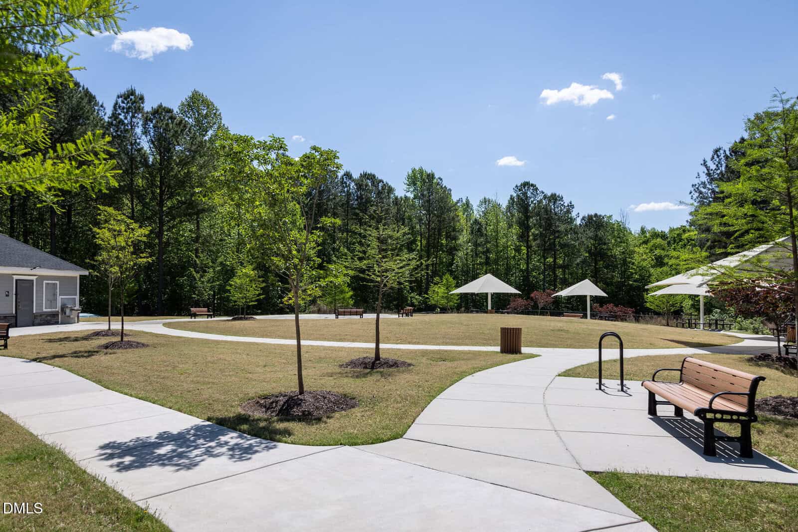 A paved walkway curves through a grassy park area with benches, young trees, and white umbrella-covered picnic tables, bordered by a forest under a clear blue sky.