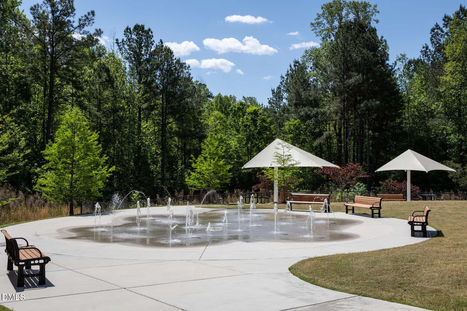 A circular splash pad with water jets, surrounded by benches and trees, with two white shade structures and a grassy area on a sunny day.