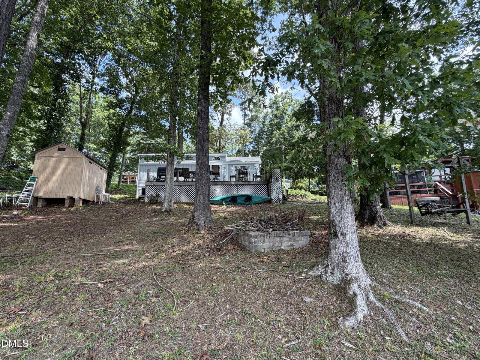 A white lake house with a porch sits among tall trees, with a small wooden shed to the left and a play structure to the right on a grassy, leaf-strewn slope.